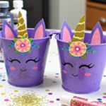 Two purple unicorn-themed mini buckets decorated with painted faces, golden horns, and pink ears, surrounded by glitter, markers, and crafting supplies on a table
