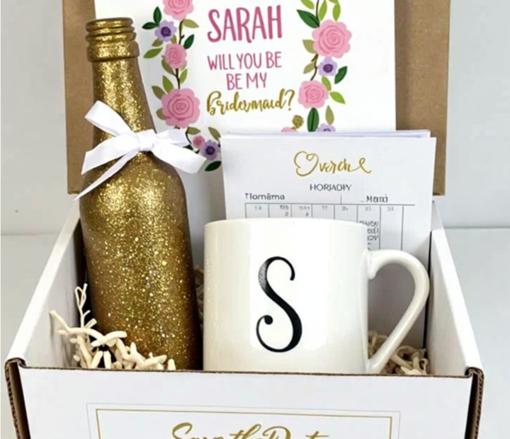 DIY bridal proposal box with a personalized bridesmaid card, a gold glitter champagne bottle, a monogrammed white mug, and a 'Save the Date' calendar, elegantly arranged in a white gift box with decorative filler