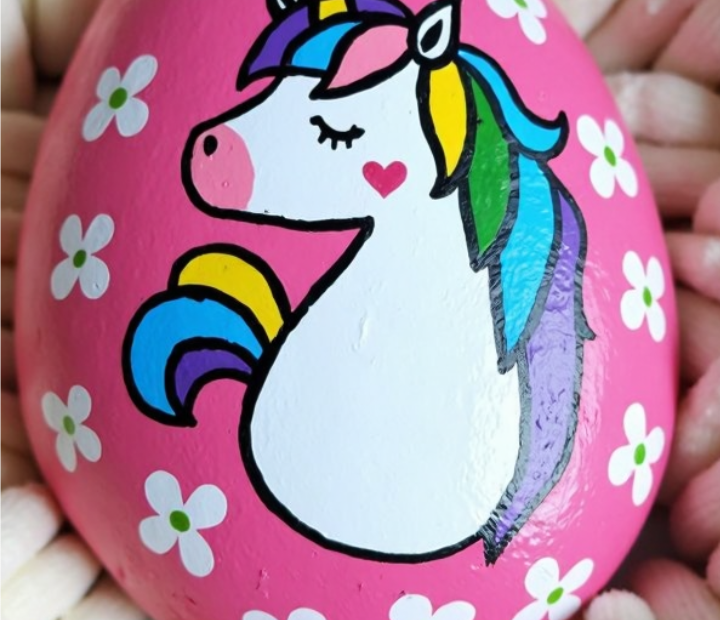 hand-painted pink rock featuring a white unicorn with a colorful rainbow mane, surrounded by small white flowers with green centers, resting on a soft cream-colored fabric