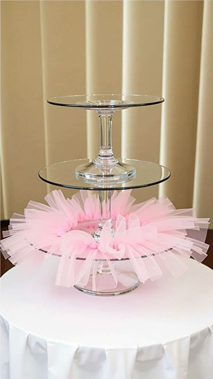 Two-tier glass cupcake stand with a pink tulle tutu decoration around the bottom tier, placed on a white table against a neutral curtain backdrop