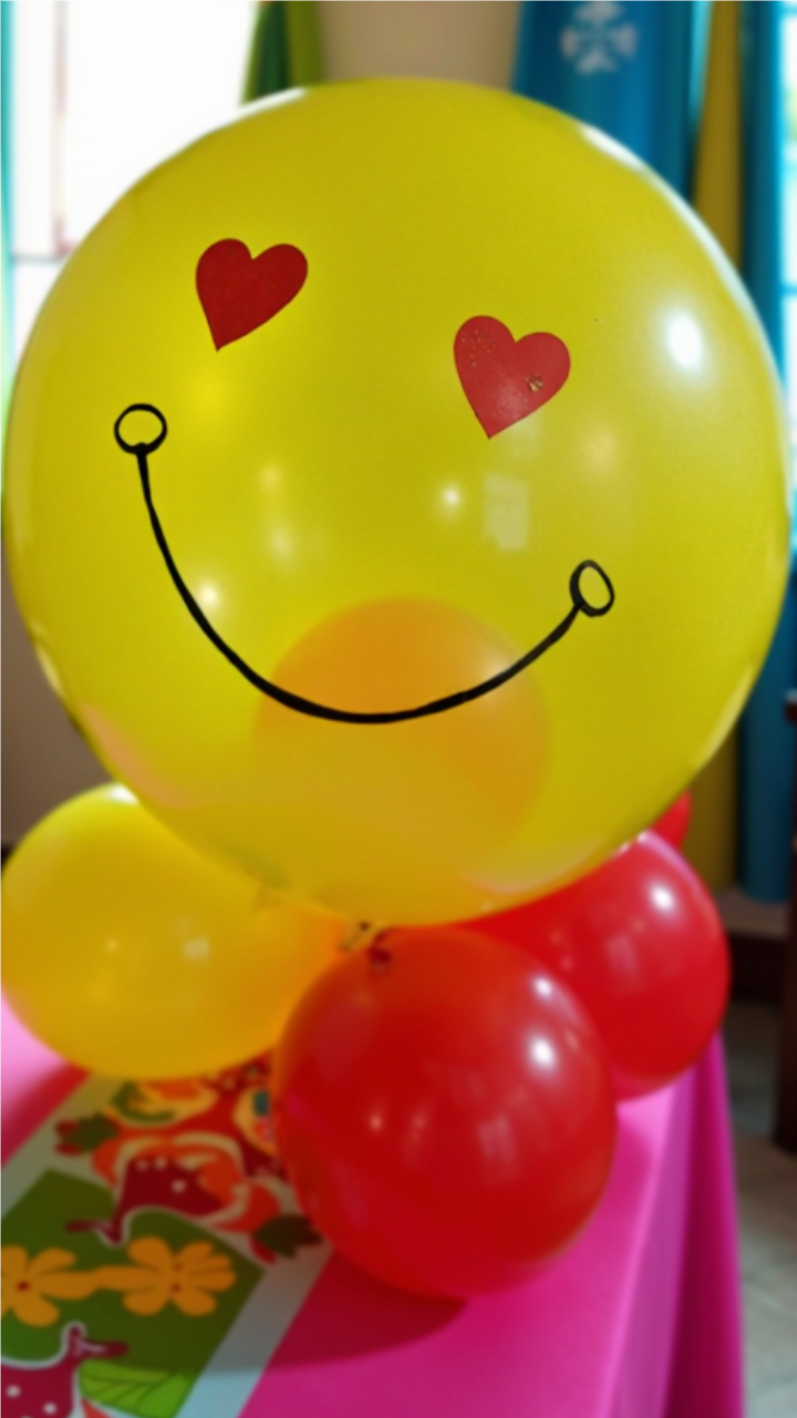 A yellow emoji balloon with heart eyes and a smiling face, surrounded by red and yellow balloons, placed on a pink party table as a fun decoration