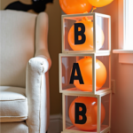 Stacked clear boxes filled with orange balloons spelling 'BABY', set against a Halloween-themed backdrop with black bat decorations