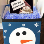 DIY Snowman Care Package gift box filled with holiday treats, decorated with a snowman face and snowflakes on a blue background