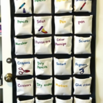 A black hanging organizer with multiple white fabric pockets, each labeled with colorful handwritten text. Some labels contain misspellings or jumbled letters of common school supplies, such as 'Tape,' 'Chalk,' 'Pencils,' 'Markers,' 'Scissors,' and 'Glue stikes.' The organizer is hung on a white door and is used for back-to-school organization