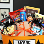 Halloween-themed movie night gift basket filled with popcorn, candy, soda, and spooky decorations like fake spiders and a spiderweb on the wall. The orange bucket has a 'Movie Night' label with black stars, and a small wrapped gift with a red ribbon sits among the treats